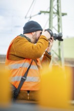 Person with camera wearing safety vest focused in industrial environment, Hermann Hesse Bahn