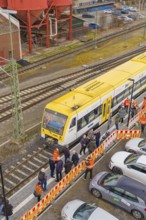 A yellow train at a train station with people and parked cars on the side, acceptance of the