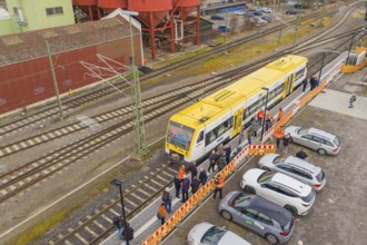 Yellow train at a train station with parked cars and people, red industrial structure, acceptance