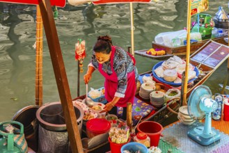 Elderly woman wearing mask offers food on her wooden boat, Damneon Saduak floating markets channel,