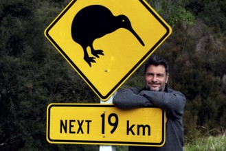 Warning sign with kiwi symbol on a road with a man in the background, Franz Josef Glacier, New