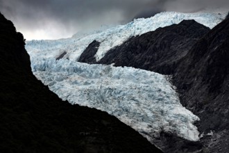 The Franz Josef Glacier contrasts strongly with dark, steep rocks under a threatening sky, Franz