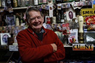 Man leans at the well-stocked bar of the Barrow Creek Hotel and smiles surrounded by souvenirs and