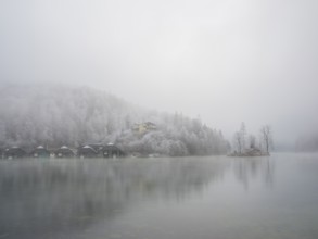 View across Königssee to boathouses, Christlieger island and frozen trees in fog, Schönau am