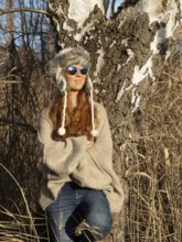 Slender pretty woman with fur hat and pompoms, red hair and warm winter colors between birch forest