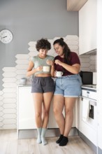 A happy lesbian couple stands in a kitchen, chatting and sharing a morning coffee. They enjoy