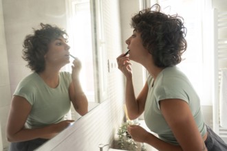 A woman with curly hair and a green top applies lipstick while looking in the mirror of a bright,