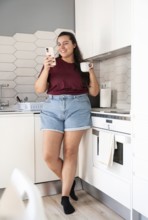 A curvy woman in casual clothes stands in a bright kitchen, holding a phone and a mug, smiling. The