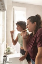 A lesbian couple shares a morning routine in the bathroom, with one applying makeup and the other