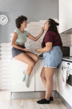 A happy lesbian couple shares a playful moment in a modern kitchen. One partner sits on the