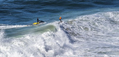 Surfers with their jet ski pilots in the Atlantic waves below Farol de Nazaré, Forte São Miguel,