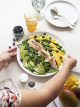 Woman placing a plate of healthy tuna, mango, avocado and blueberry salad on a table