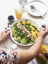 Woman seasoning a colorful healthy salad with chicken, mango, avocado, blueberries and lamb's