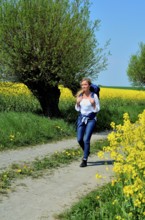 Young woman with backpack walking on a countryroad with willow trees and field of rape in Skivarp,