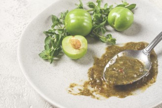 A spoon with tkemali sauce, on a plate, with cherry plum and mint, top view, no people