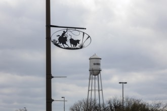 Bandera water tower behind artistic metal sign showing cowboy life on pole in Bandera, Texas, USA
