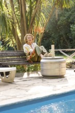 Elderly woman enjoys a moment of peace, sitting on a bench in her garden near the swimming pool,