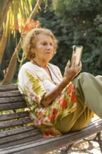 Elderly woman enjoying a tranquil moment in the park, sitting comfortably on a bench while browsing