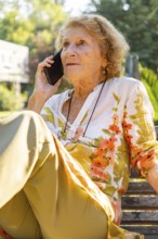 Senior woman sitting comfortably on a park bench, making a phone call and enjoying the warm summer