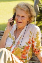 Elderly woman with glasses hanging from a string around her neck is sitting on a wooden bench