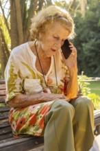 Senior woman sitting on a wooden bench in a serene garden, using her smartphone to enjoy