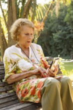 Senior woman sitting on a wooden bench in a park, enjoying the pleasant weather while using her