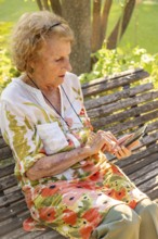 Senior woman sitting on a wooden bench in a park, enjoying the warmth of summer while using her