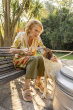 Senior woman sitting on a wooden bench in a sunny garden, interacting with her pet dog, creating a