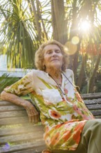 Elderly woman with closed eyes enjoying sunlight while sitting on wooden bench in lush botanical