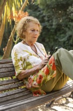 Senior woman sitting comfortably on a wooden bench in a park, using a tablet while enjoying the