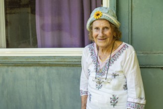 Elderly woman smiling while leaning against a light green wooden door, wearing a light blue hat