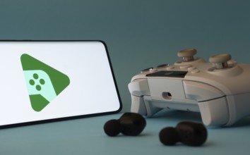 A smartphone with a Google Play Games app icon next to a controller and earbuds on a blue surface