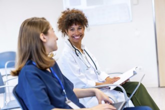 Two female doctors are sitting and smiling while discussing medical information during a conference