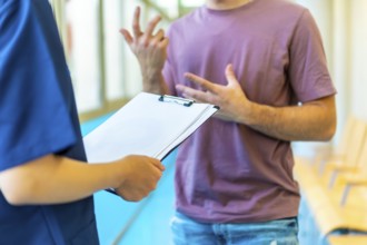 Young man explaining his symptoms using gestures to a nurse holding a medical chart in a hospital