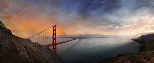 Panoramic view of the Golden Gate Bridge with a rainbow at sunset and orange-glowing storm clouds,
