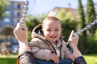 Smiling baby boy enjoying a swing ride pushed by his mother at the playground on a sunny day