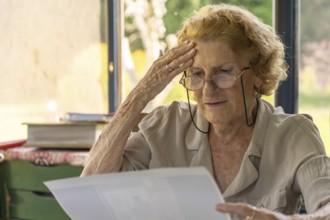 Senior woman wearing glasses, reading a letter at home and feeling a wave of worry, touching her