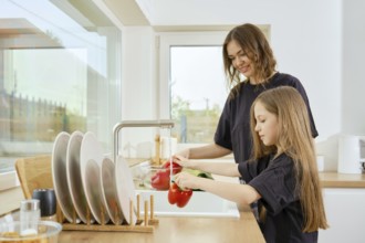 A mother watches her daughter wash vegetables under running water in the kitchen