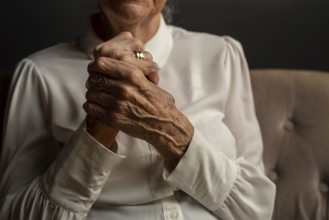 90 year old woman in white blouse holding folded hands, conveying thoughtfulness in a dim light,