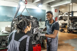 Two smiling mechanics are working on a car engine suspended by a chain hoist in a professional