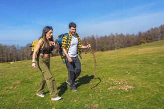 Young couple walking hand in hand across a vibrant green meadow, carrying backpacks and using a