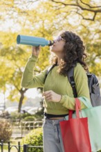 Woman with curly hair and sunglasses drinks from a blue water bottle in a park. She carries