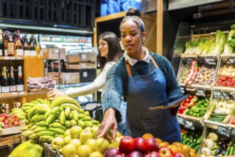 Supermarket employee using digital tablet while arranging fresh fruit on display, customer choosing