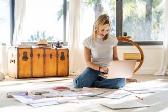 A woman sits on a bright home floor, absorbed in her laptop, with papers scattered around her.