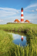 The Westerheversand lighthouse under a blue sky with white clouds reflected in a tideway,