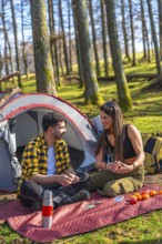 Couple playing cards near tent and backpack in forest during camping trip, enjoying sunny day in