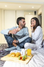 Young couple wearing pajamas sitting in bed eating donuts for breakfast, with croissants, orange