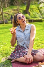 Cheerful young woman laughing while eating pizza in a vibrant green park, enjoying a sunny spring