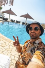 Tourist man in the hotel pool in summer taking a selfie enjoying the vacation, making the victory