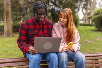 Two happy young students are using a laptop and a smartphone while sitting on a bench in a park,